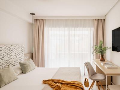 Lichte hotelkamer met comfortabel bed, bureau, stoelen en groot raam met gordijnen.
