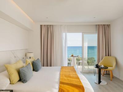 Modern hotelkamer met tweepersoonsbed, gele accenten en zeezicht door grote ramen.