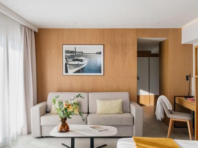 Modern hotelkamer met bank, houten muur, bureau en groot raam met lichte gordijn.
