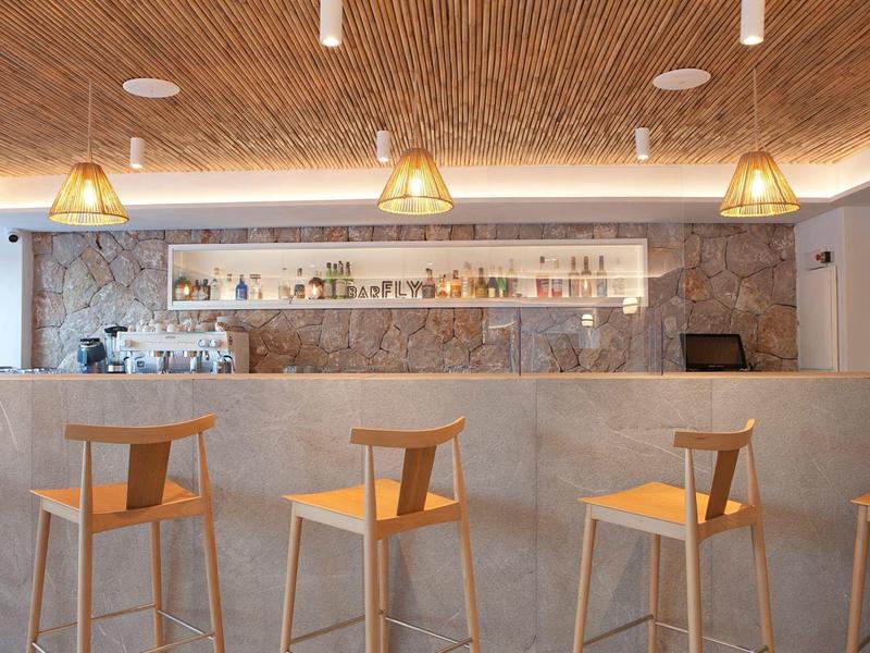 Nowoczesne wnętrze baru z drewnianymi stołkami, kamienną ścianą i ciepłym oświetleniem sufitowym.