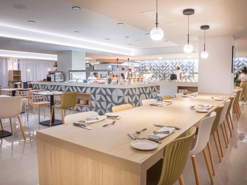 Nowoczesna restauracja z długimi drewnianymi stołami, białymi krzesłami i oświetlonym barem.