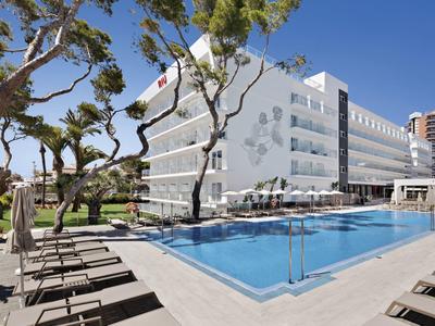 Moderner Hotelkomplex mit rechteckigem Pool, Liegestühlen und schlanken Bäumen bei blauem Himmel.