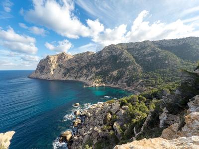 Küstenlandschaft mit felsigen Klippen, blauem Meer und bewaldeten Hügeln unter bewölktem Himmel.