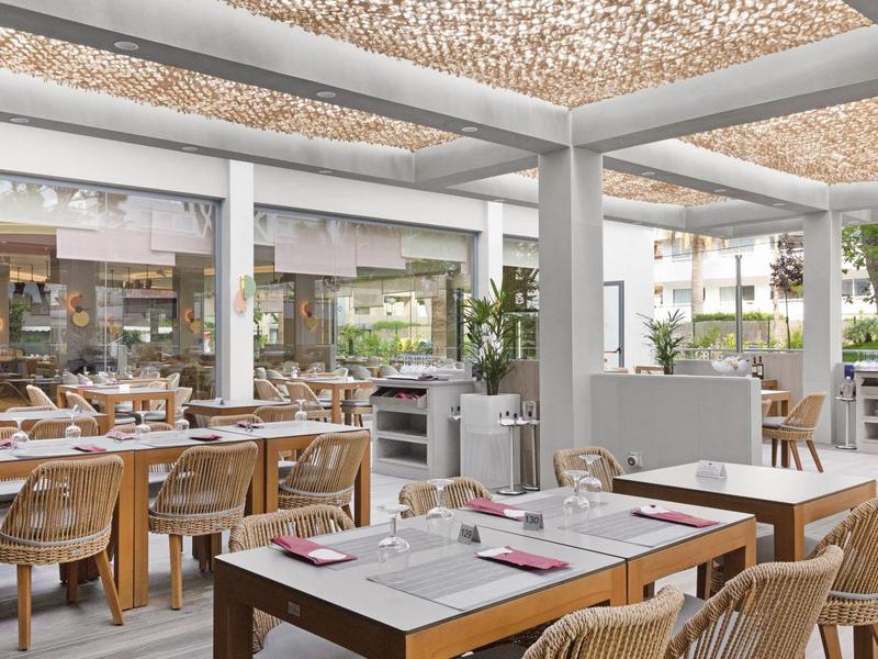 Moderner Außenbereich eines Restaurants mit Holztischen, geflochtenen Stühlen und Sonnenschutz.