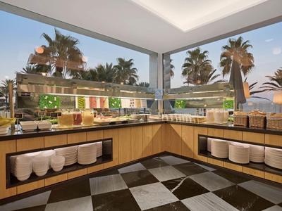 Moderní snídaňový koutek s výhledem na palmy za soumraku, vybavený různým jídlem a teplým osvětlením.