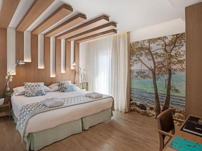 Světý hotelový pokoj s manželskou postelí, dřevěnými akcenty a nástěnným obrazem lesa u moře.