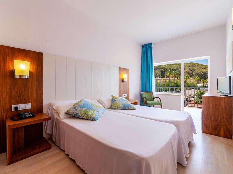 Chambre d'hôtel lumineuse avec deux lits simples et balcon offrant une vue sur des collines verdoyantes.