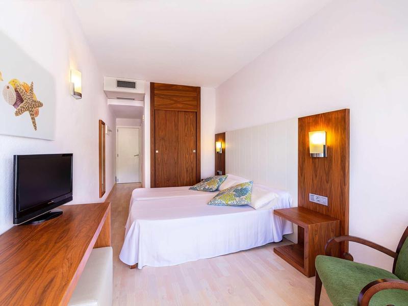 Chambre d'hôtel simple avec deux lits simples, TV, meubles en bois et fauteuil vert.