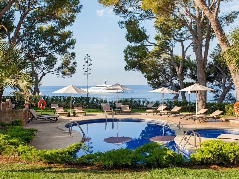 Runder Pool mit blauem Wasser, Liegestühlen und Schirmen am Meer unter Bäumen bei Sonnenschein.