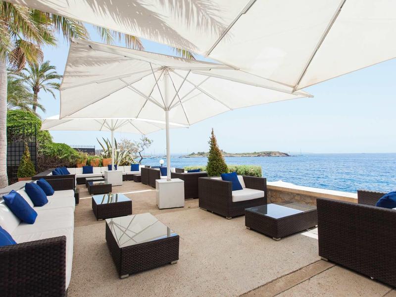 Terrassenlounge mit weißen Sonnenschirmen, dunklem Sitzmobiliar und blau-weißen Kissen am Meer.