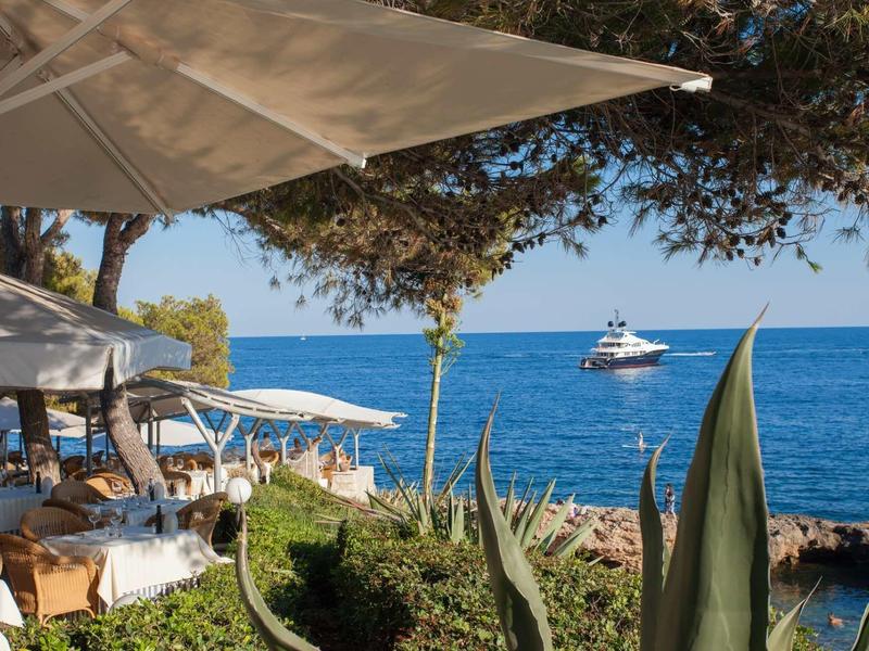 Strandrestaurant mit Sonnenschirmen, Palmen und einem Boot auf dem blauen Meer im Hintergrund.
