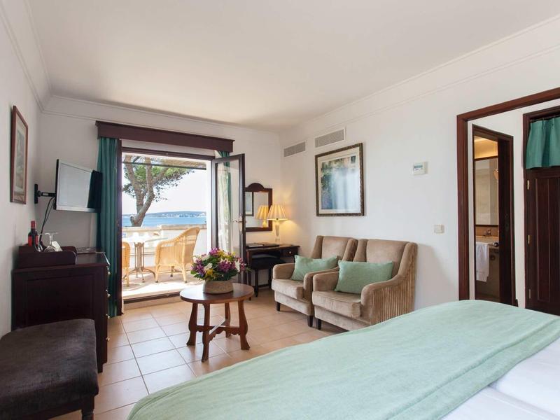 Helles Schlafzimmer mit hellgrünem Bett, Sofa, Holzmöbeln und Blick auf Terrasse und Meer.