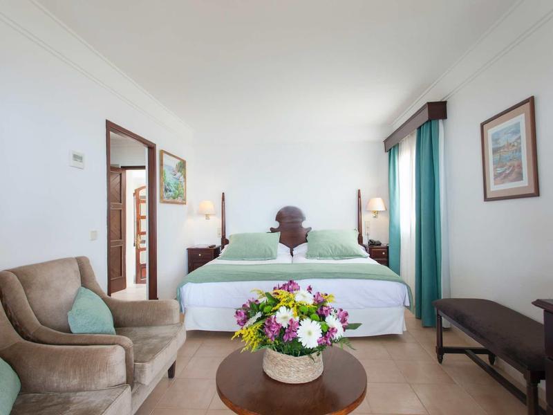 Helles Schlafzimmer mit großem Bett, bequemen Sessel, rundem Tisch mit Blumen und grünen Vorhängen.