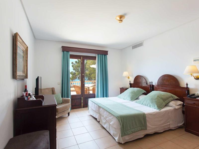 Hell eingerichtetes Schlafzimmer mit Doppelbett, Holzmöbeln, Fliesenboden und Balkontür mit Vorhängen.