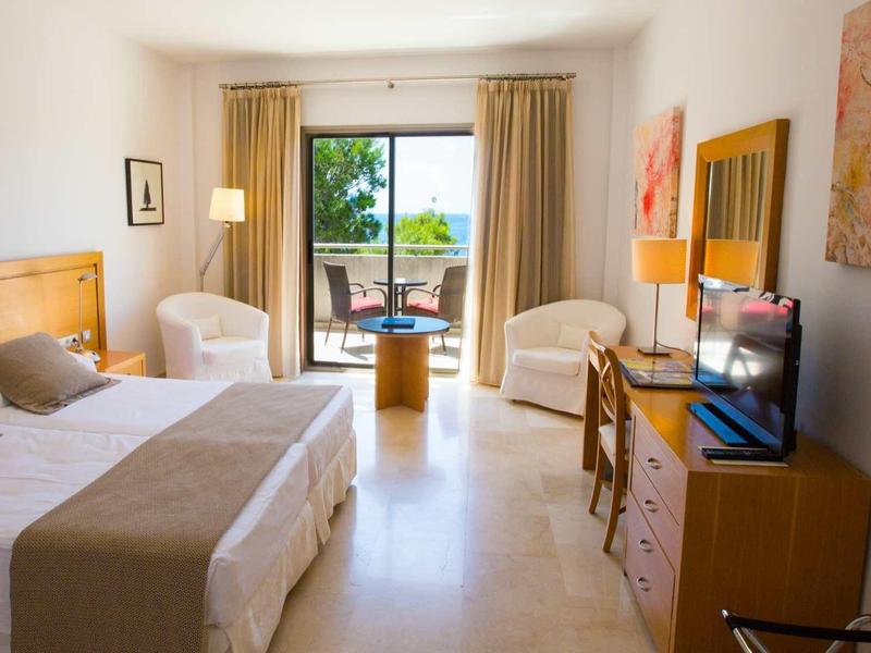 Helles Doppelzimmer mit hellen Möbeln, zwei Sesseln, einem Schreibtisch und Blick auf Terrasse mit Meer.