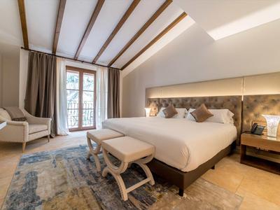 Jasno urządzona sypialnia z dużym łóżkiem, krzesłami i dywanem przy oknie z zasłonami.