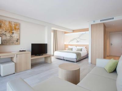 Nowoczesny pokój hotelowy z podwójnym łóżkiem, sofą, telewizorem i biurkiem w jasnych kolorach.