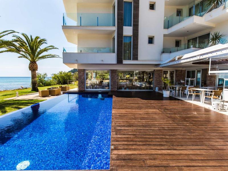 Nowoczesny basen hotelowy z drewnianym tarasem, widokiem na morze i palmami przy słonecznej pogodzie.