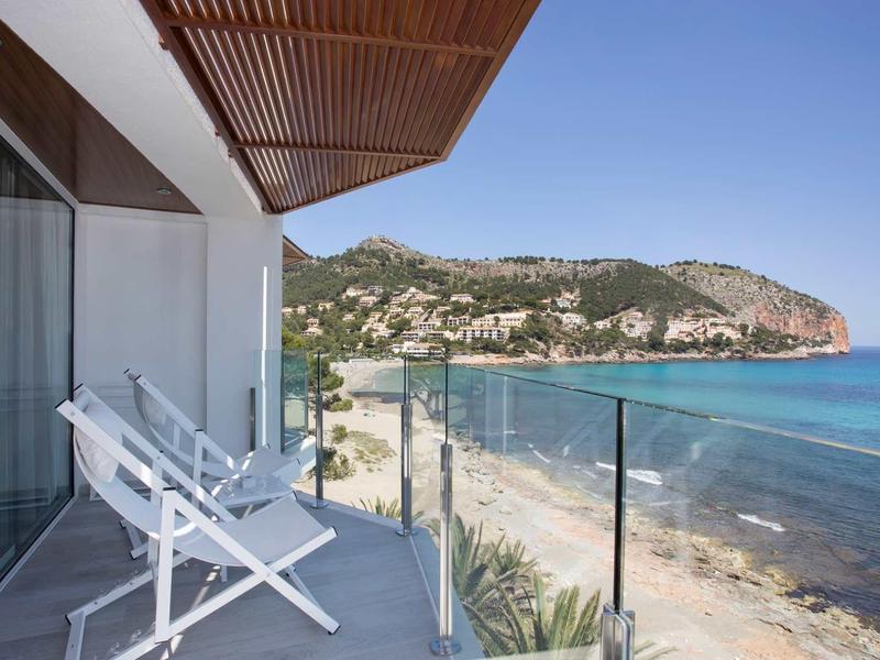 Balkon z białymi leżakami i szklaną barierką z widokiem na morze i odległe klify.