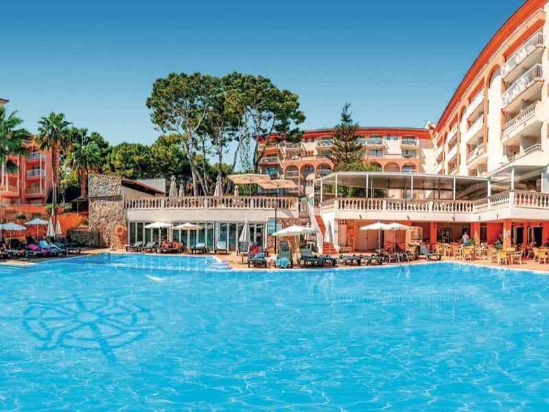 Grand piscine d'hôtel avec parasols et bâtiments à plusieurs étages en arrière-plan sous un ciel clair.