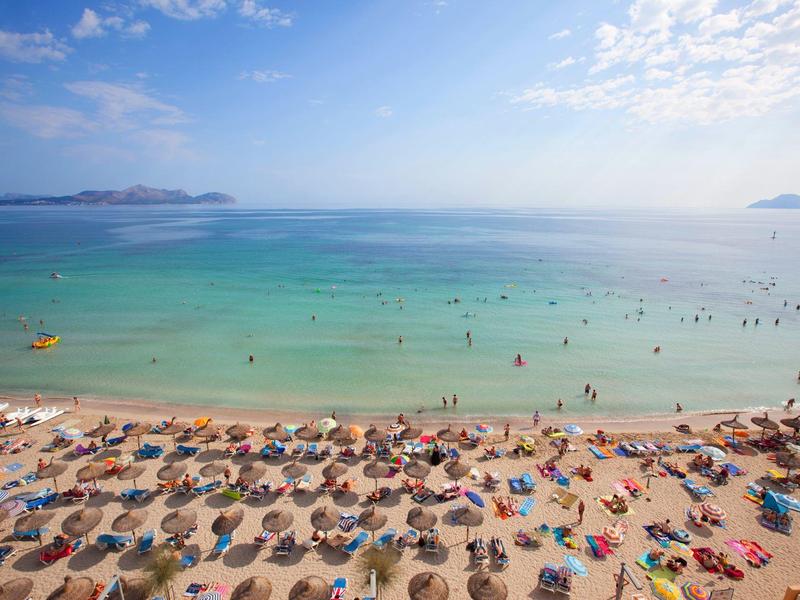 Spiaggia sabbiosa affollata con molti ombrelloni e persone, cielo azzurro e mare limpido.