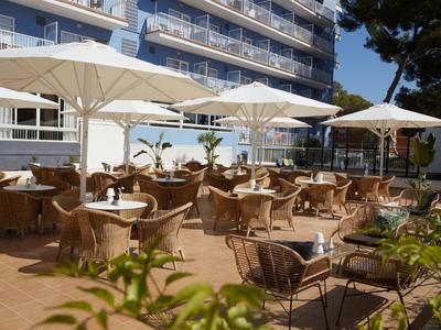 Freiluftrestaurant mit weißen Sonnenschirmen und geflochtenen Stühlen vor einem Hotelgebäude.