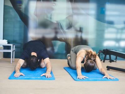 Zwei Personen machen Dehnübungen auf blauen Yogamatten in einem hellen Raum.