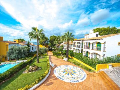 Ośrodek z białymi budynkami, palmami i centralną brukowaną ścieżką pod słonecznym niebem.