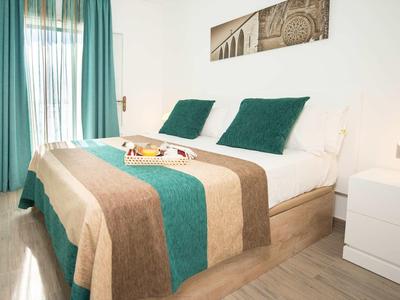 Przytulna sypialnia z podwójnym łóżkiem, zielono-brązową narzutą i jasnymi zasłonami.