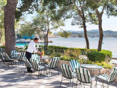 Terrasse mit Tischen und Stühlen neben Wasser und Bäumen, Kellner serviert Getränke