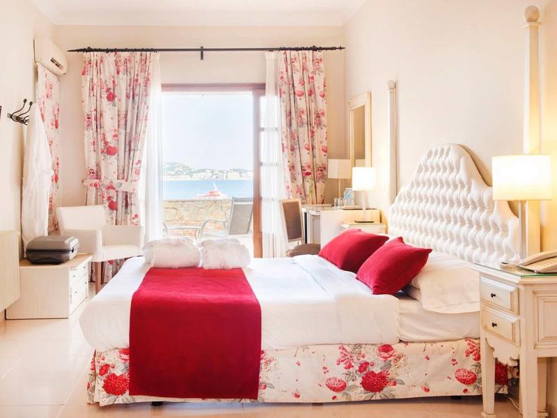 Jasny pokój hotelowy z widokiem na morze i kwiecianą pościelą.