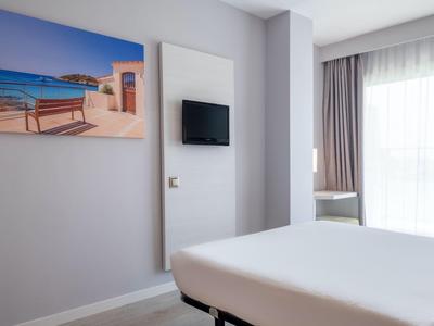 Nowoczesny pokój hotelowy z dużym łóżkiem, oknem z zasłonami i obrazem morza na ścianie.