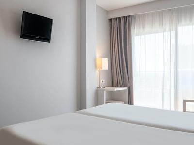 Jasny pokój hotelowy z dwoma łóżkami, telewizorem na ścianie i lampą obok okna balkonowego.