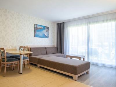 Jasny pokój hotelowy z sofą, stołem jadalnym, krzesłami i dużym oknem z zasłonami.