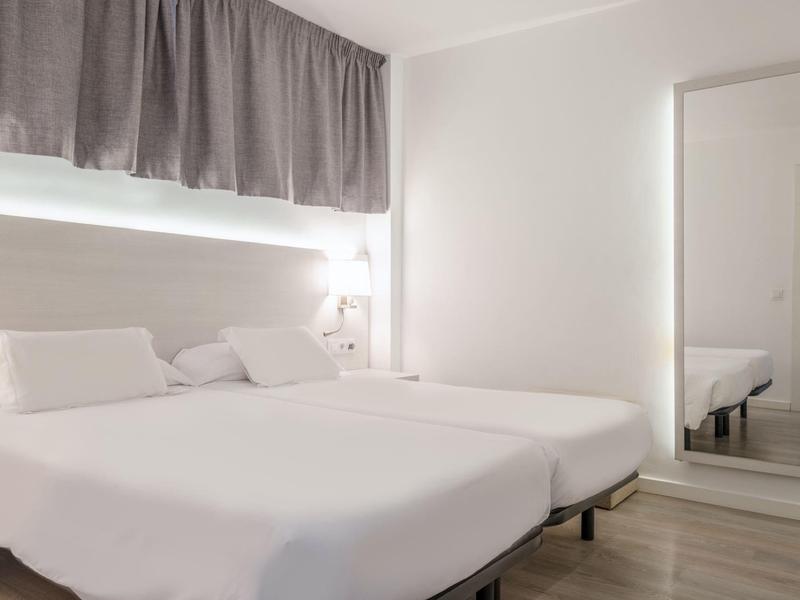 Jasny pokój hotelowy z dwoma pojedynczymi łóżkami, dużym lustrem i zasłoną nad oknem.