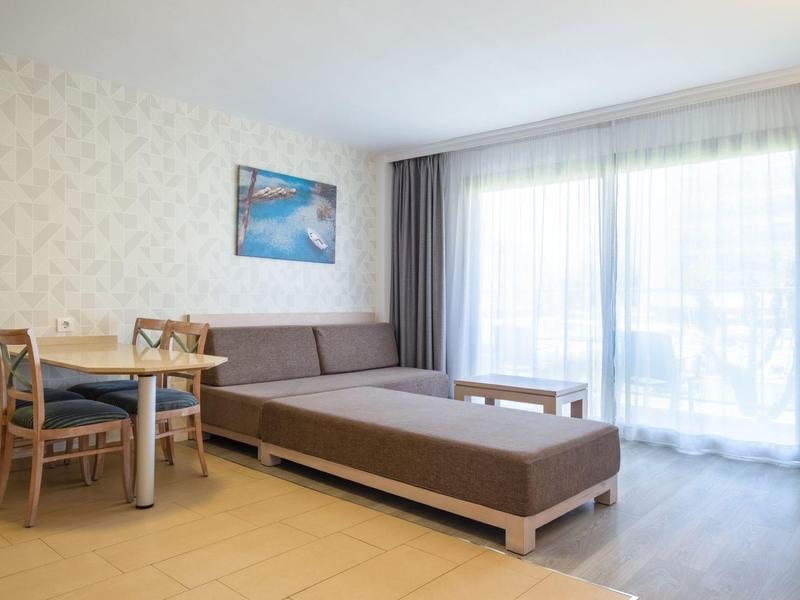 Jasny pokój hotelowy z sofą, stołem jadalnym, krzesłami i dużym oknem z zasłonami.