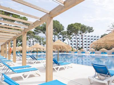 Großer Pool mit blauen Liegestühlen und Strohdächern neben Holzpergola und Bäumen im Hintergrund.