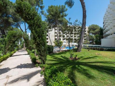 Grüner Hotelgarten mit Sonnenschirmen, Pool und weißen Gebäuden unter blauem Himmel.