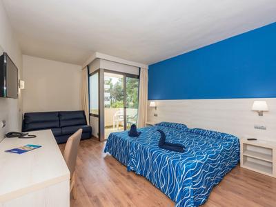 Moderní hotelový pokoj s modrou postelí, pohovkou, psacím stolem a balkonem s výhledem do zeleně.