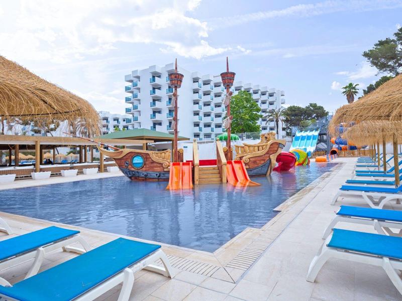 Dětský bazén s herními prvky, obklopený modrými lehátky a střešními střechami v hotelu.