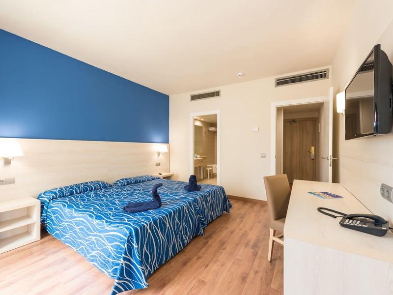 Moderní hotelový pokoj s modrou akcentní zdí, manželskou postelí, psacím stolem a televizí.