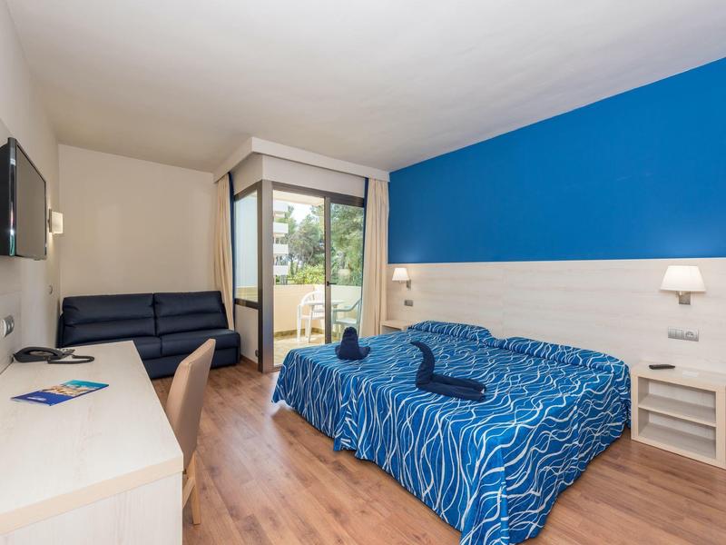 Moderní hotelový pokoj s modrou postelí, pohovkou, psacím stolem a balkonem s výhledem do zeleně.