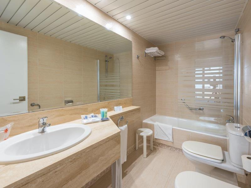 Moderní koupelna s umyvadlem, vanou, toaletou a velkými zrcadly v světlých odstínech.