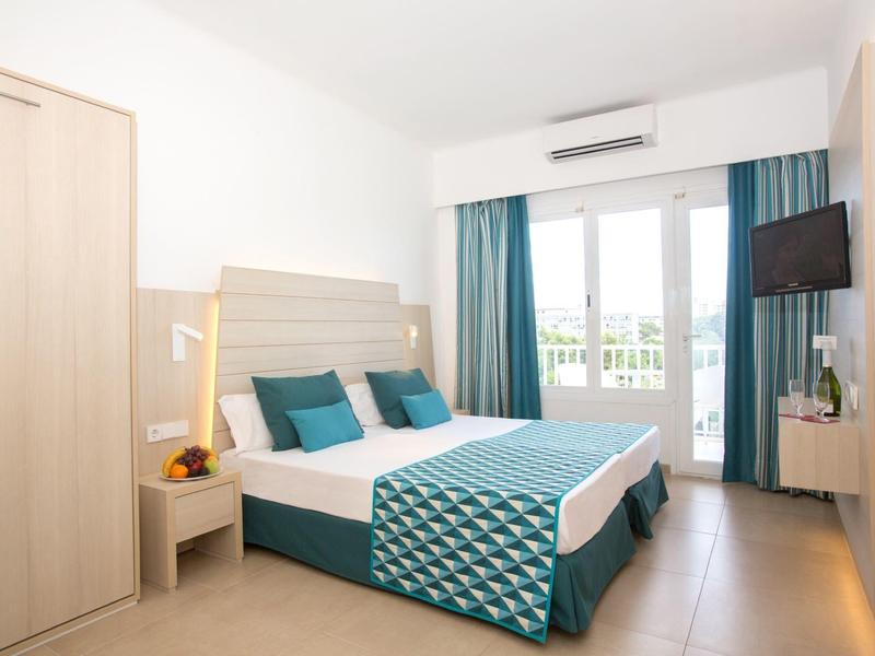 Chambre d'hôtel moderne avec lit double, literie bleu-vert, télévision et balcon avec porte en verre.