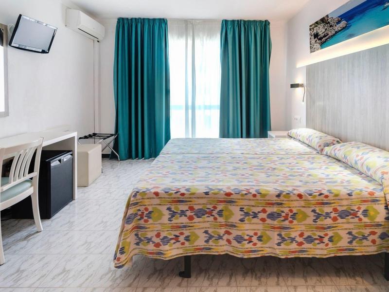 Helles, modernes Hotelzimmer mit Doppelbett, buntem Bettbezug, Schreibtisch, TV und türkisfarbenen Vorhängen.