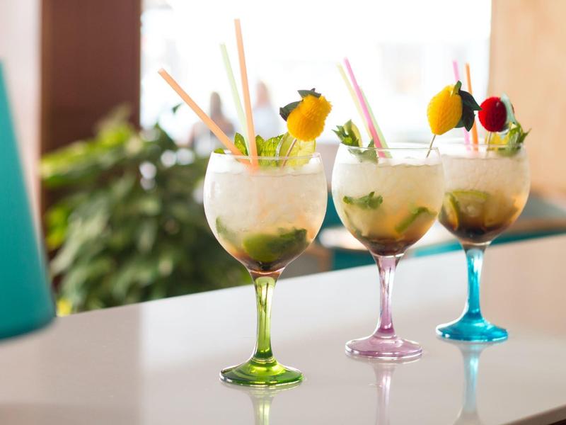Three colorful cocktails in large glasses with fruit and straws on a white counter.