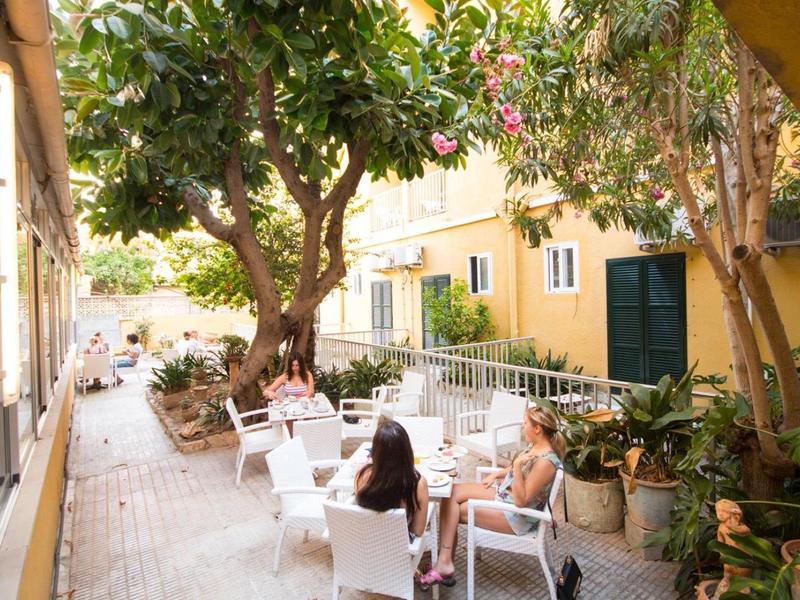 Cozy outdoor area with white chairs and tables under green trees beside yellow buildings.