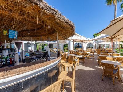 Außenbereich einer Hotelbar mit Strohdach, Tischen und Stühlen unter Sonnenschirmen.