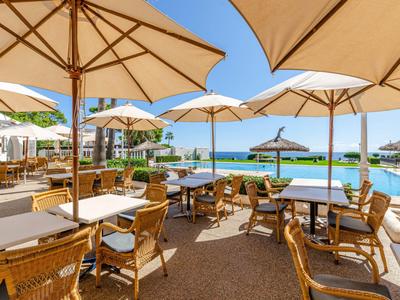 Außenbereich eines Hotels mit Sonnenschirmen, Tischen und Stühlen neben einem Pool bei klarem Himmel.
