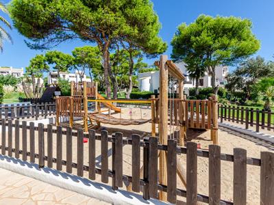 Spielplatz mit Klettergeräten und Sandboden, umgeben von einem Holzzaun und Bäumen neben einem Hotel.